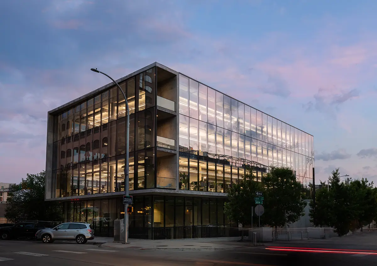 A modern glass building at twilight, reflecting a pink and blue sky. Lit interior reveals office spaces. Cars are parked on the street, conveying a calm ambiance.