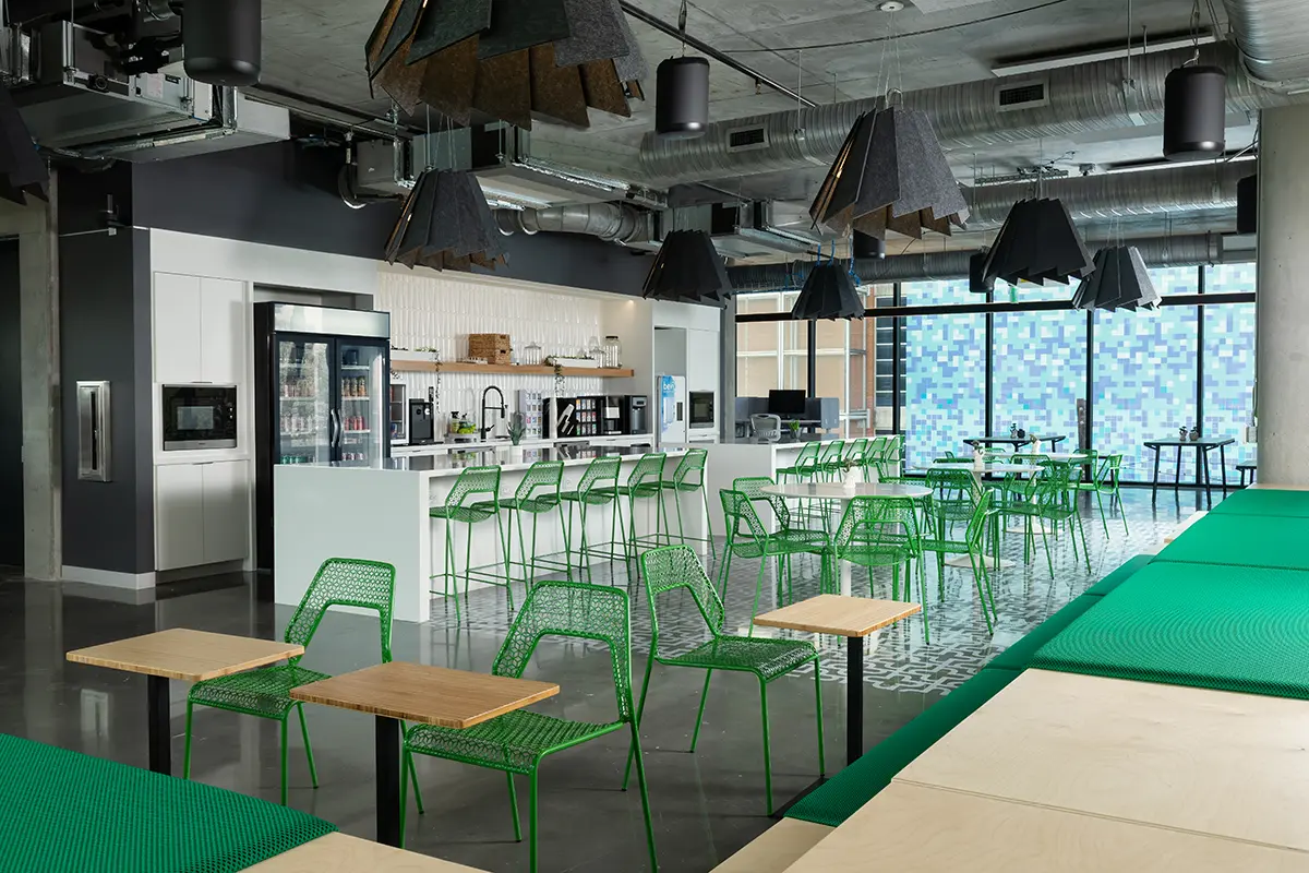 Modern office cafeteria with green wire chairs, wooden tables, and geometric ceiling lights. Large windows reveal a pixelated blue pattern.