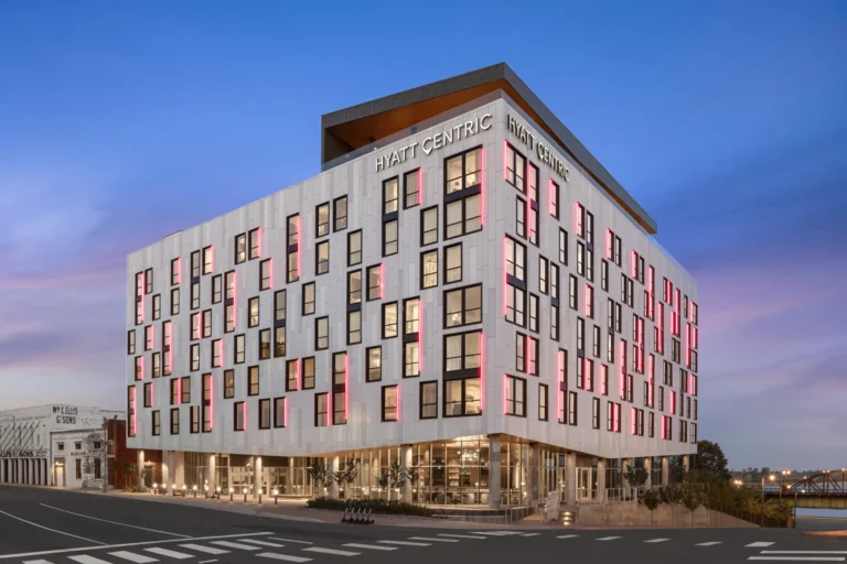 Modern hotel exterior at sunset, featuring a white facade with geometric black-framed windows. Pink lighting accents create a vibrant, welcoming ambiance.