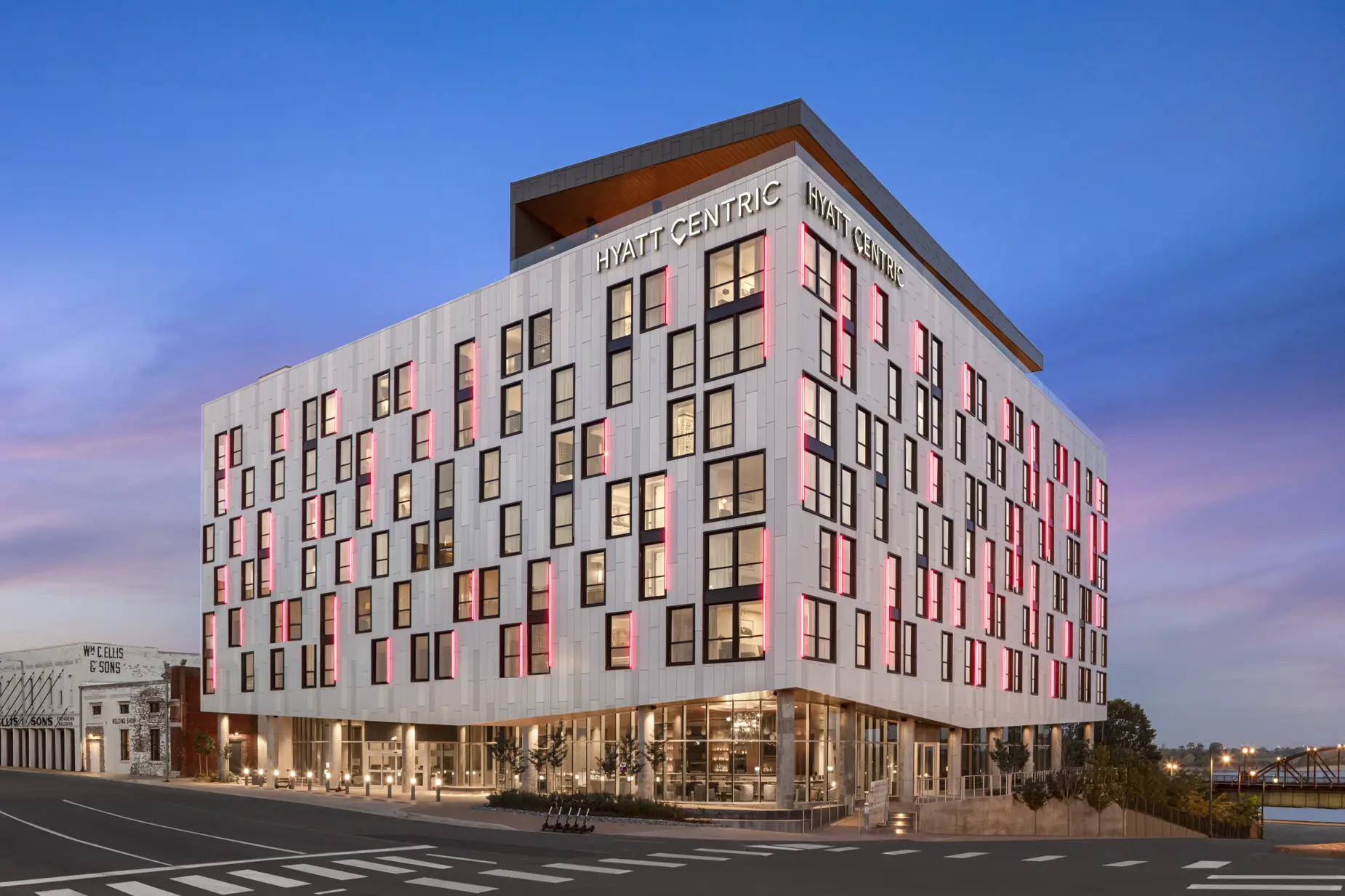 Modern hotel exterior at sunset, featuring a white facade with geometric black-framed windows. Pink lighting accents create a vibrant, welcoming ambiance.