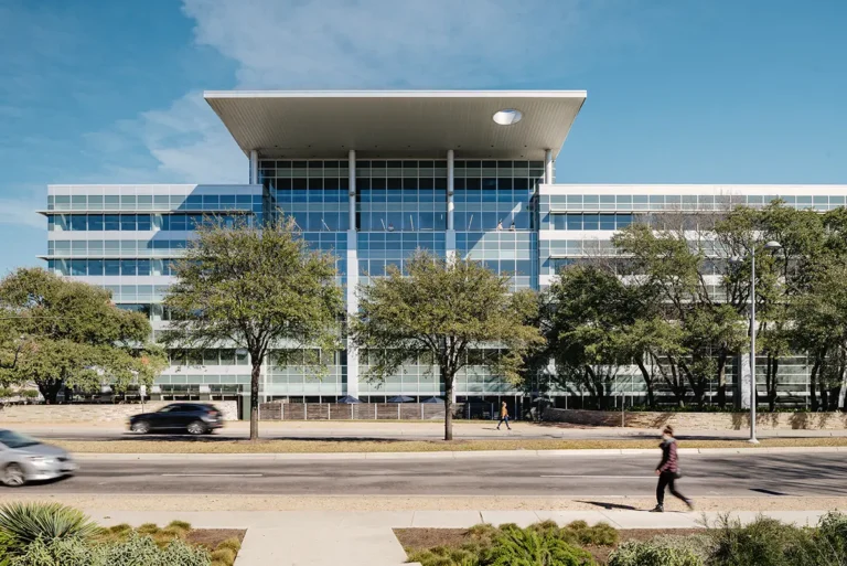 A modern glass building with large windows stands behind trees on a sunny day. A car drives by, and a person walks along the sidewalk in the foreground.