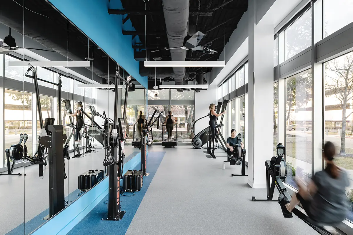 Modern gym interior with large windows and mirrored walls. Several people use equipment, including rowing machines and step climbers. Bright and energetic atmosphere.