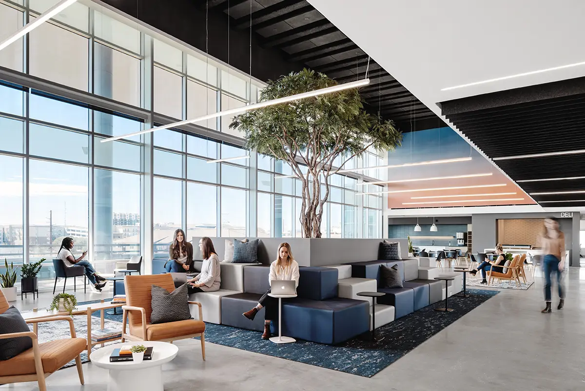 Modern office lounge with large windows, modular seating, and a central indoor tree. People work on laptops, creating a calm, collaborative vibe.