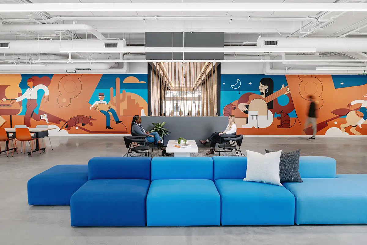 A modern office lounge with a vibrant mural depicting diverse activities lines the wall. Bright blue sectional couch and two people conversing add a lively, creative vibe.