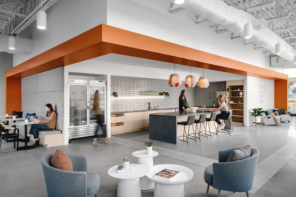 Modern office kitchen and lounge with sleek furniture, warm lighting, and a vibrant orange ceiling. People are casually interacting and working.