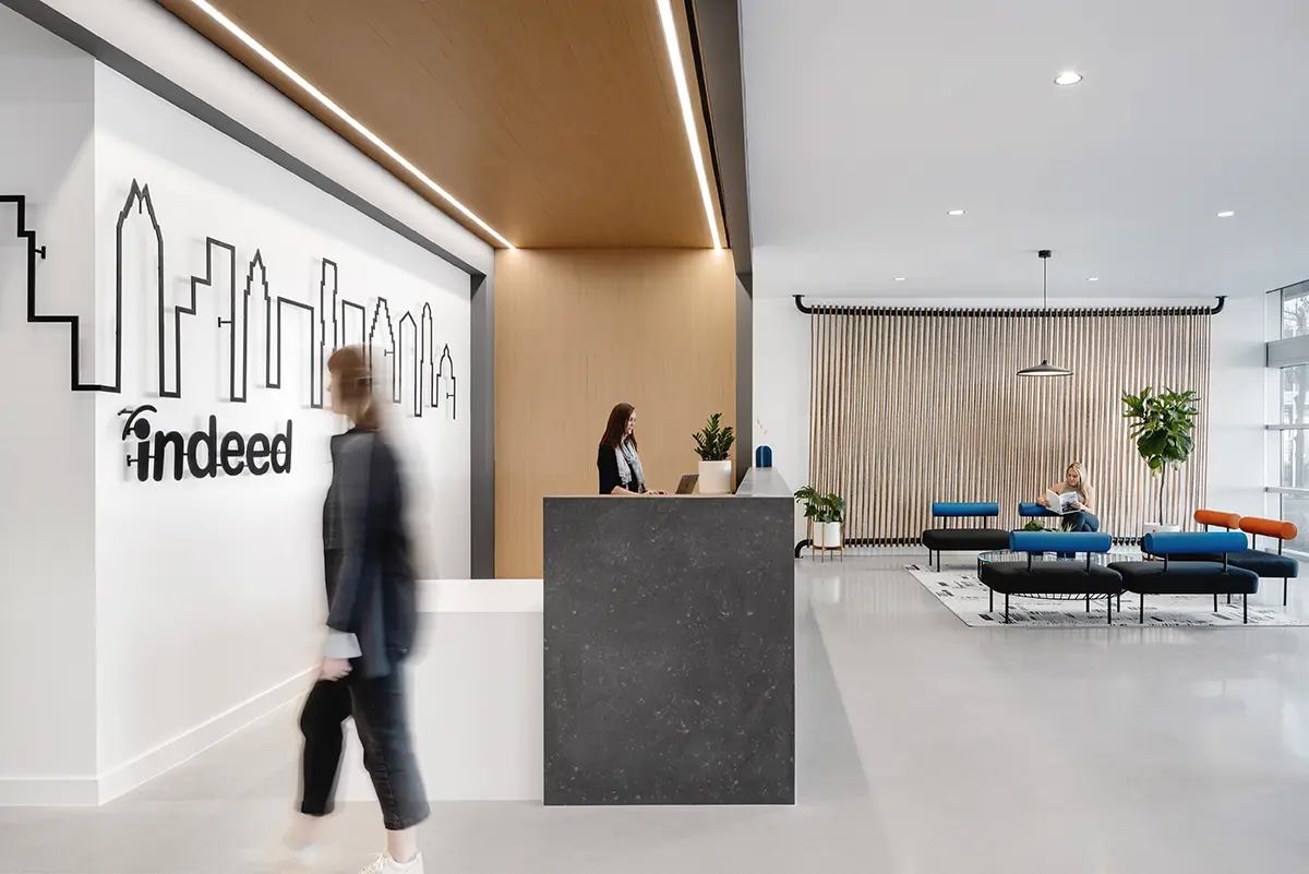 Modern office lobby with a minimalist design, featuring a reception desk and a wall with a city skyline outline. A person walks by as another sits on colorful lounge chairs, creating a welcoming and contemporary environment.