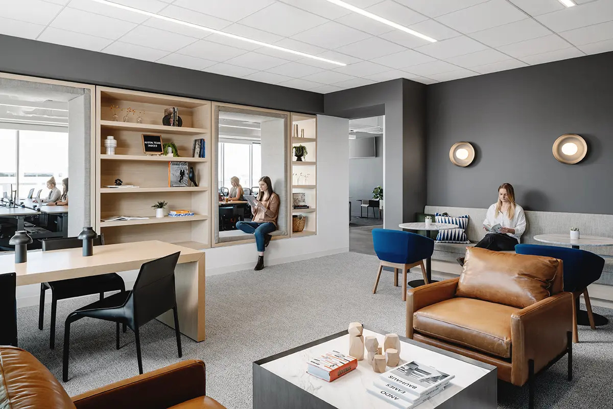 A modern office lounge with gray walls features cozy leather chairs, a bookcase, and round wall lights. People are casually seated, reading and working.