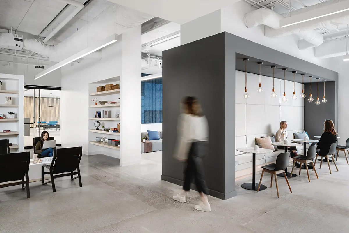 Modern open-plan office with sleek decor, featuring people working at tables and a person walking. Bright lighting and minimalistic design create a calm atmosphere.