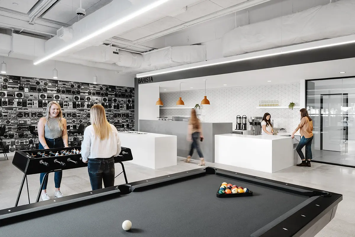 Modern office lounge with foosball and pool tables in the foreground. Two women play foosball, while others chat at a sleek barista counter.
