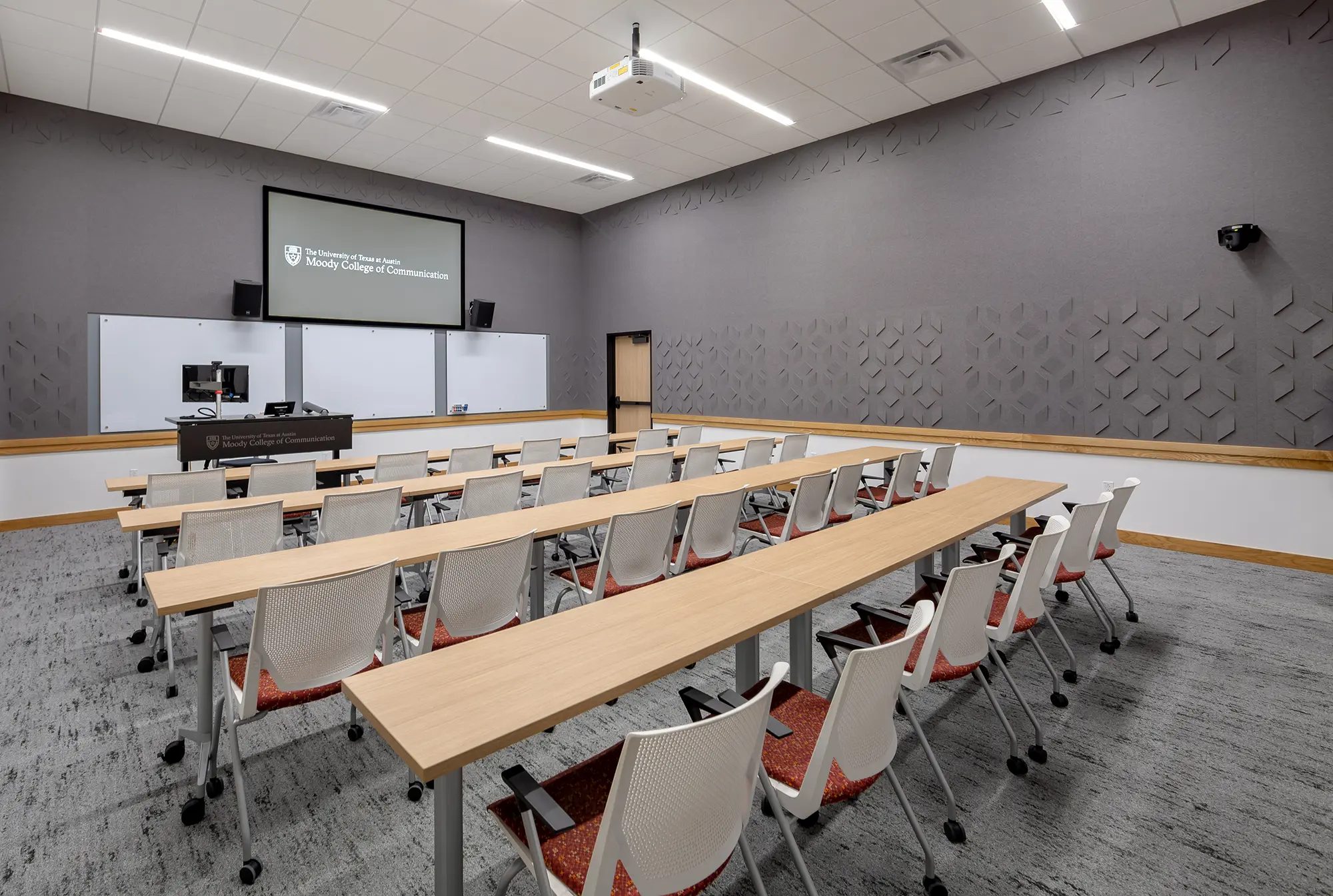 Spacious lecture room with rows of tables and white chairs facing a large screen. Modern design, grey walls, and a ceiling projector create a professional atmosphere.