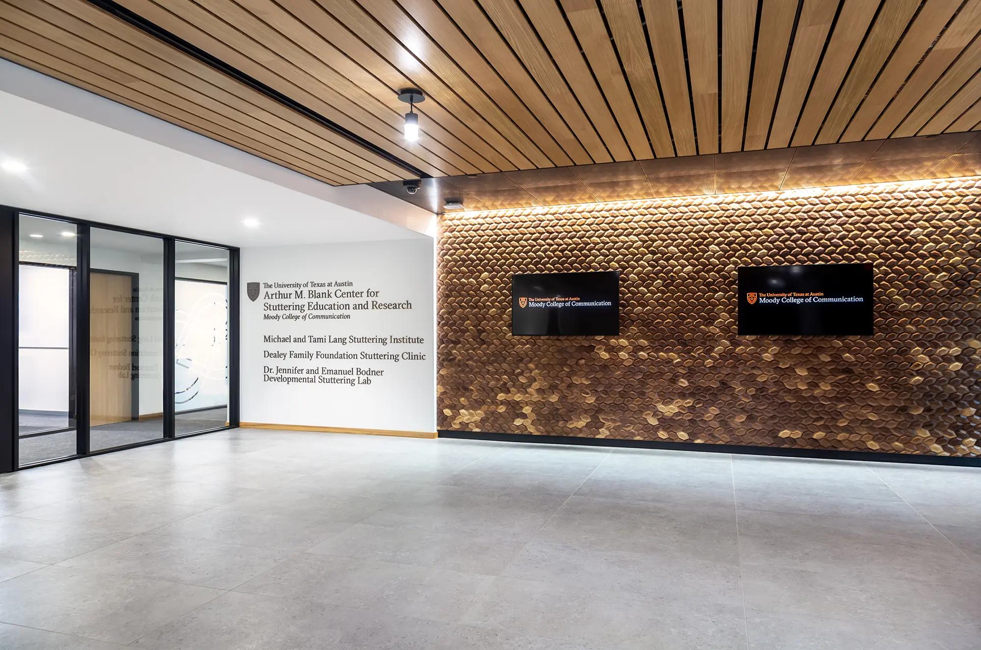 Modern interior with a textured wooden wall, featuring two screens displaying logos. A sign on the left wall indicates a stuttering research center.