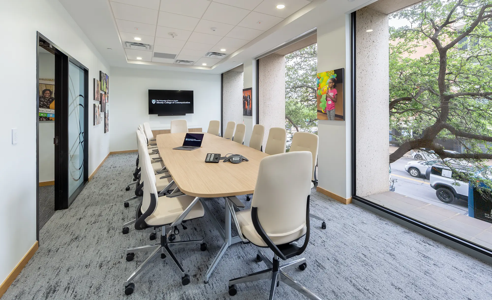 Modern conference room with a long oval table and white swivel chairs. Large windows reveal a city street and trees outside, creating an open, inviting atmosphere.
