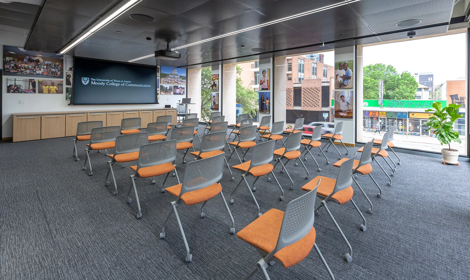 A modern lecture room with rows of gray chairs featuring orange seats. A large screen displays the Moody College of Communication logo. Windows reveal an urban view outside.