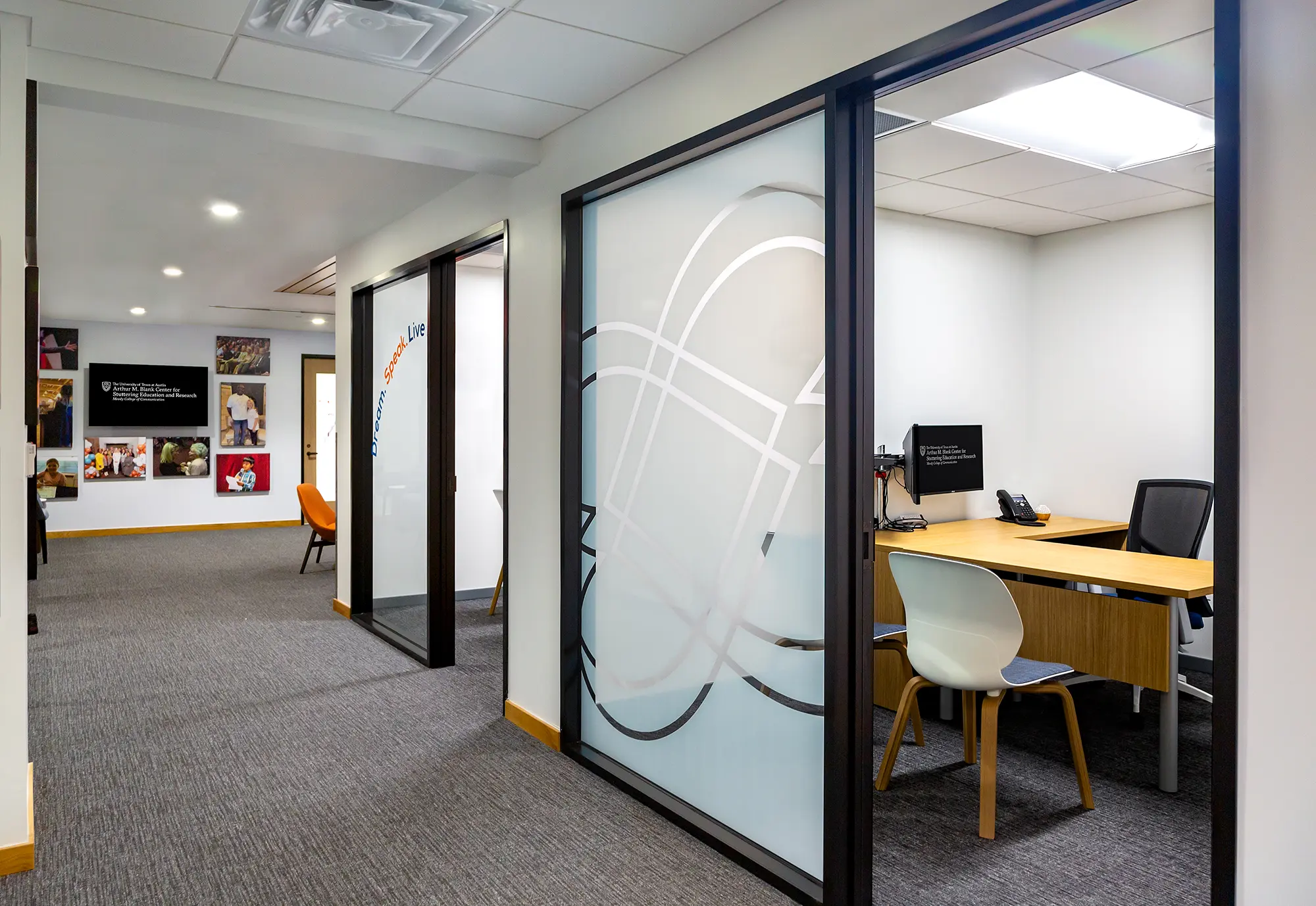 Modern office hallway with gray carpet, featuring frosted glass-walled rooms on the right. Chairs and a desk are visible inside one room. Walls display colorful artwork.