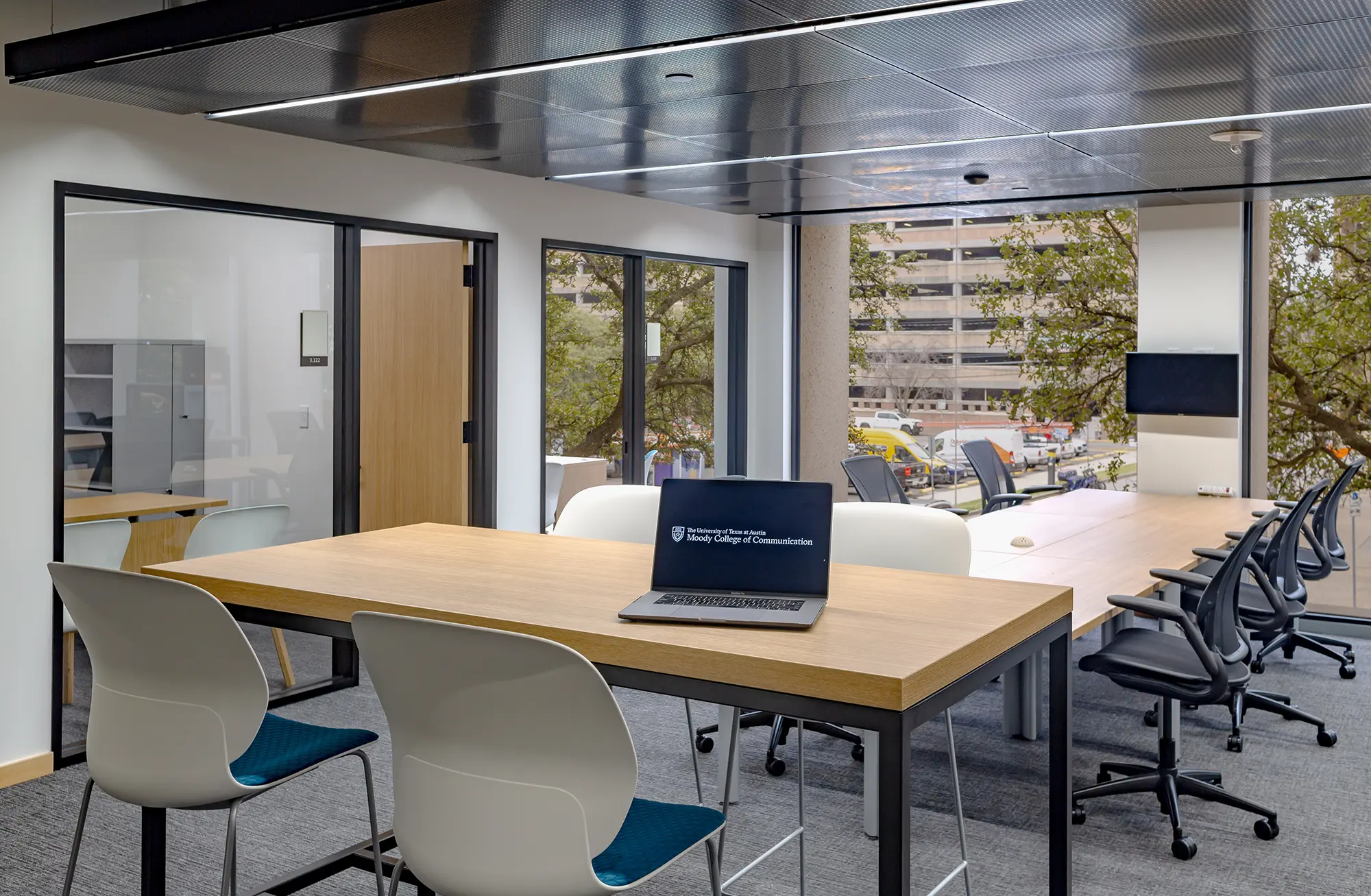 Modern office space with large windows, featuring a wooden conference table, chairs, and a laptop. Natural light creates an open, professional tone.