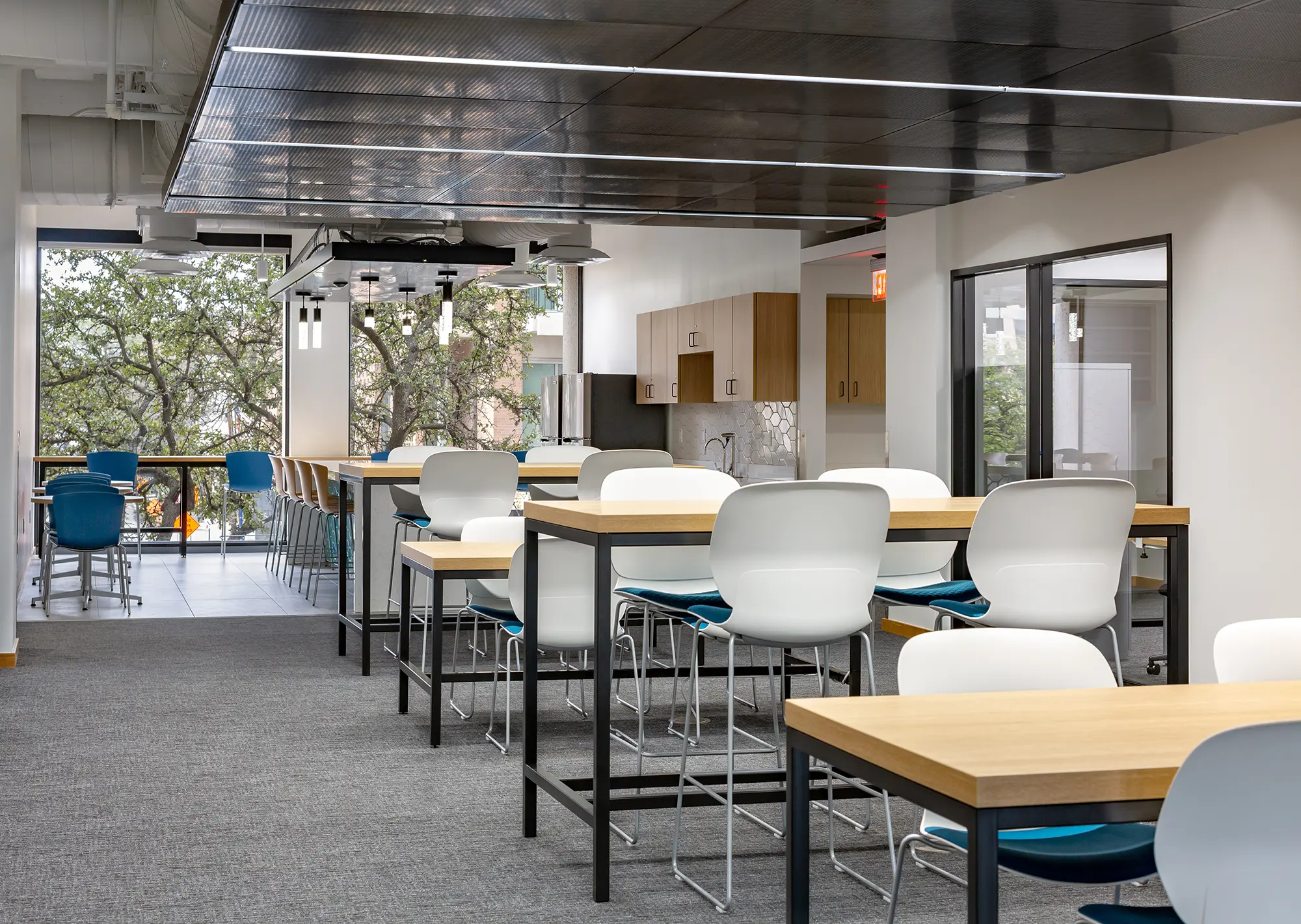 Modern office break room with tall wooden tables, white and blue chairs, and a kitchenette. Large windows reveal trees outside, creating a fresh, open vibe.