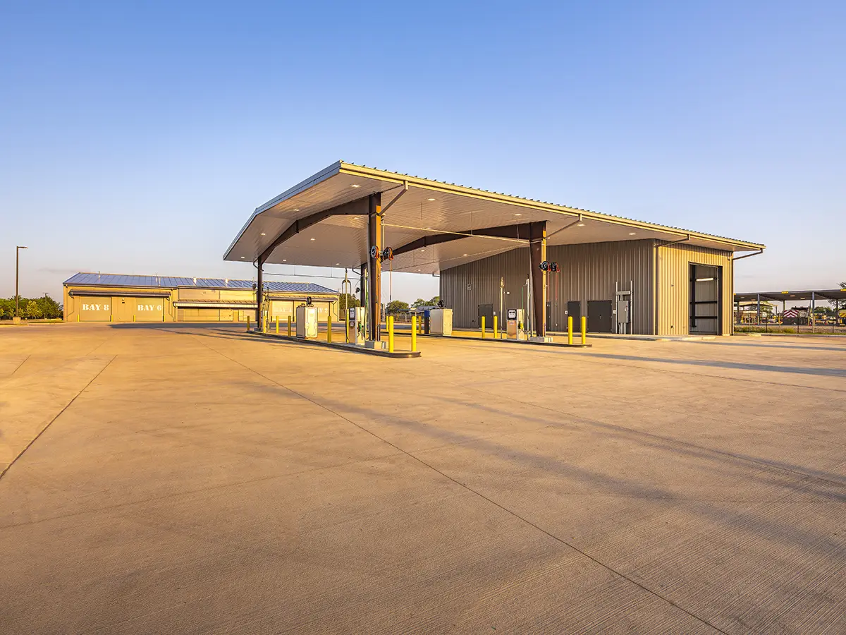 A modern, empty gas station under a broad, blue sky at sunset. Features a large canopy, fueling pumps, and a clean concrete lot, evoking a calm, spacious setting.