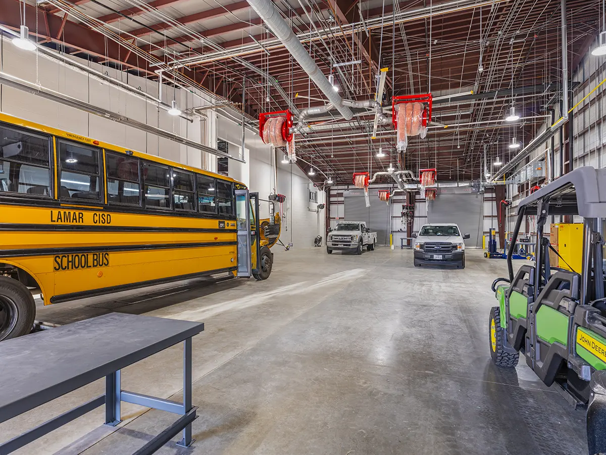 Spacious garage with yellow school bus, two pickup trucks, and green utility vehicle. High ceiling has metal pipes and red equipment. Industrial mood.