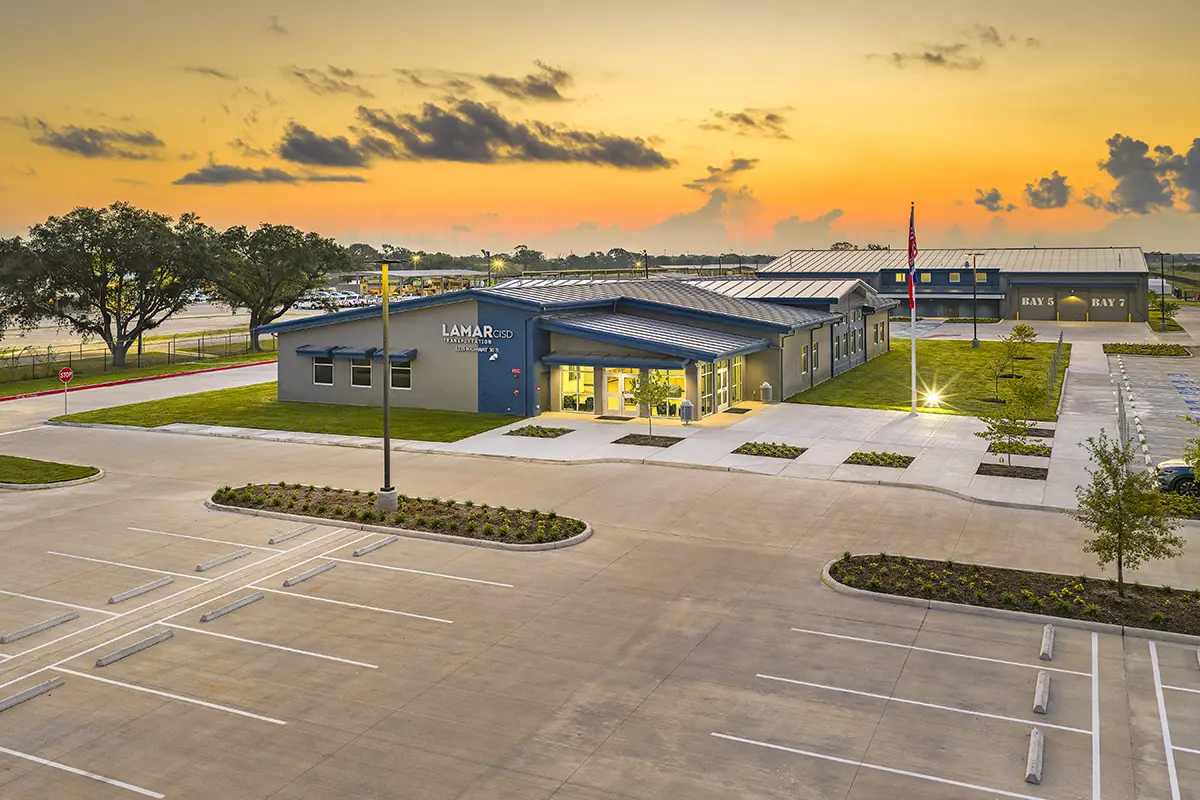 Modern building with blue accents, surrounded by a large parking lot at sunset. Trees and a flagpole are visible, creating a serene, welcoming atmosphere.