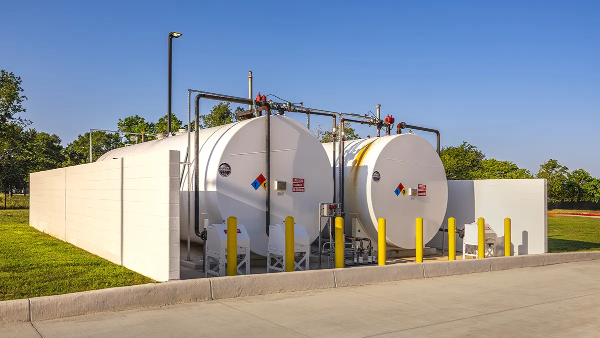 Two large white fuel storage tanks are enclosed by a low wall, with safety signs visible. The scene is set outdoors on a clear, sunny day.