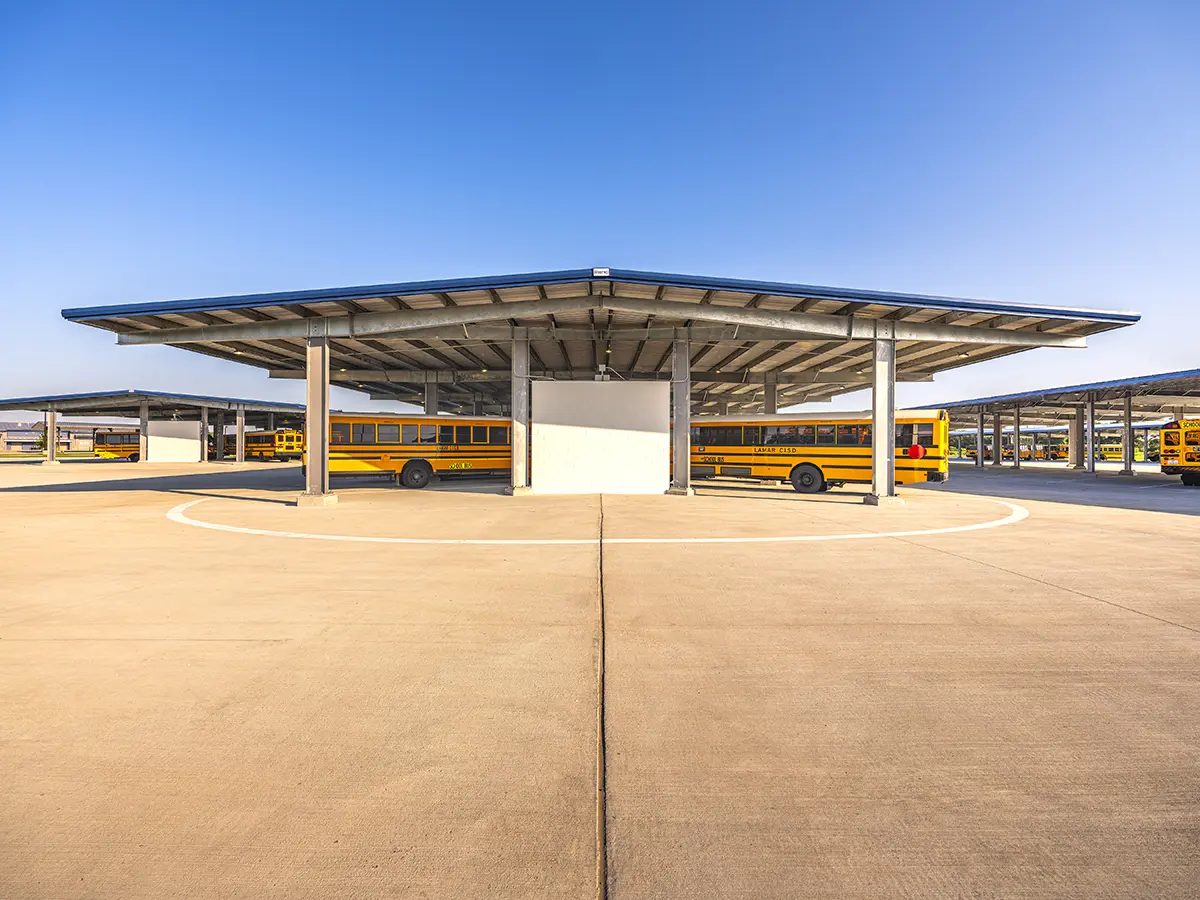 A large bus station with yellow school buses parked under expansive metal canopies. Clear blue sky and a spacious concrete lot create an orderly, calm atmosphere.