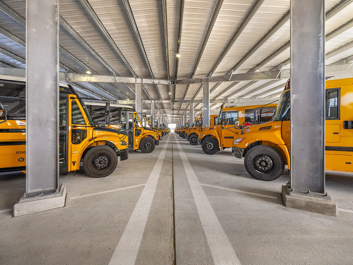 A row of yellow school buses is parked under a spacious, industrial-style garage with metal beams. The scene is orderly, bright, and expansive.