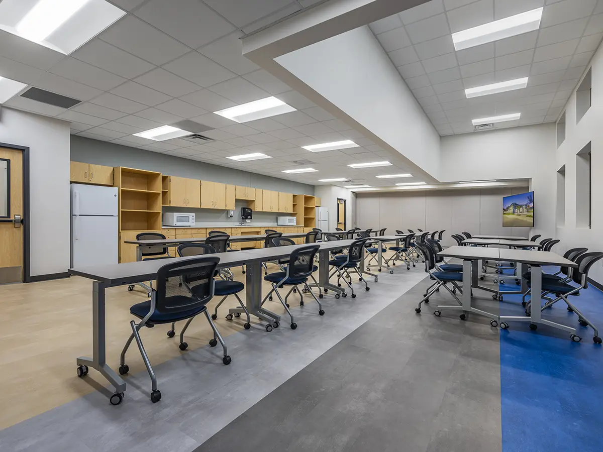 Modern classroom with long tables, rolling chairs, and a blue accent floor. Wooden cabinets hold a microwave and refrigerator. Bright lighting creates a clean, organized atmosphere.