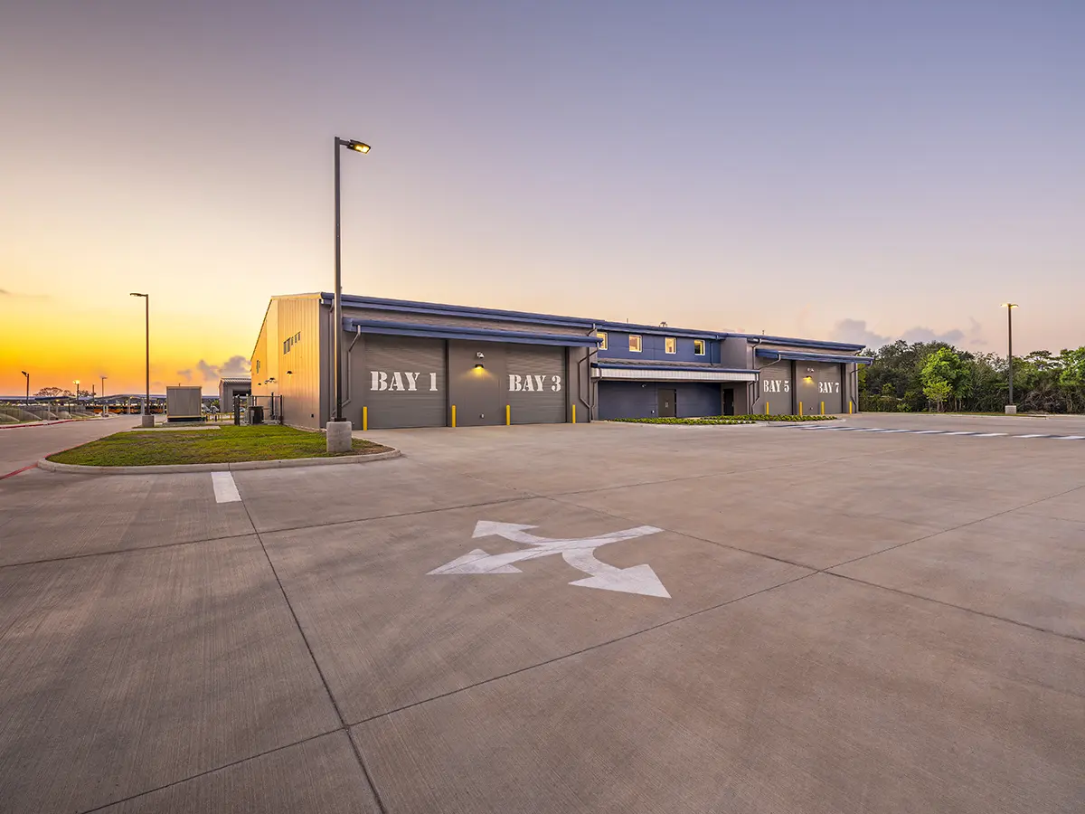Industrial building at sunset, featuring labeled bays and a spacious concrete lot with large arrows. The sky is softly glowing, adding a serene ambiance.