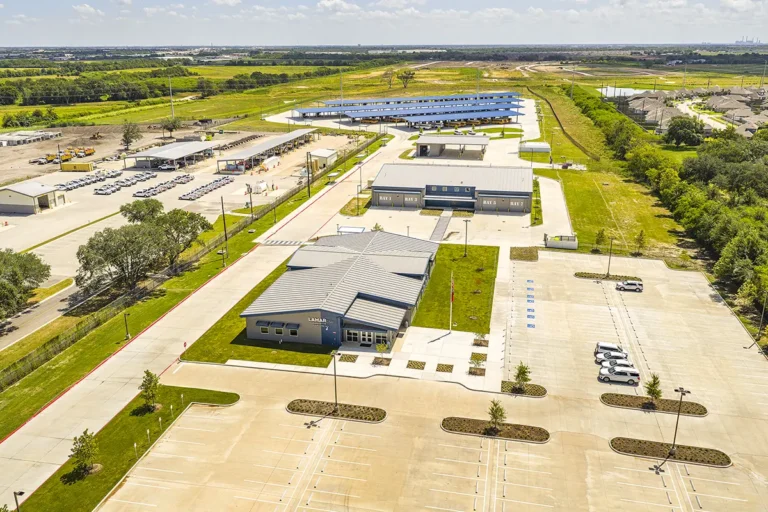 Aerial view of a large, modern driving test facility with multiple buildings and parking lots. Surrounding green fields and distant roads under a clear sky.