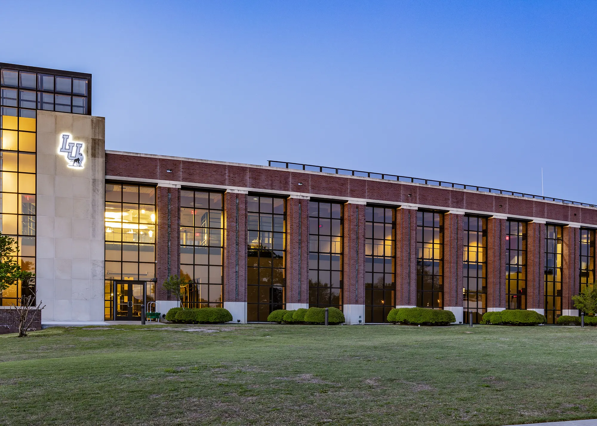 Red brick building with large reflective windows, illuminated warmly from within at dusk. A lighted logo is on the left. Calm and welcoming ambiance.