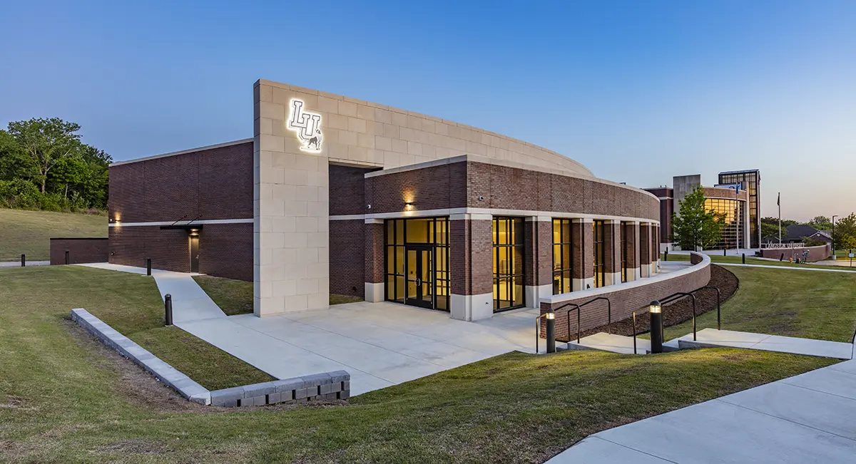 Modern brick building with large glass windows and a prominent logo on the side, surrounded by green grass and lit warmly at twilight.