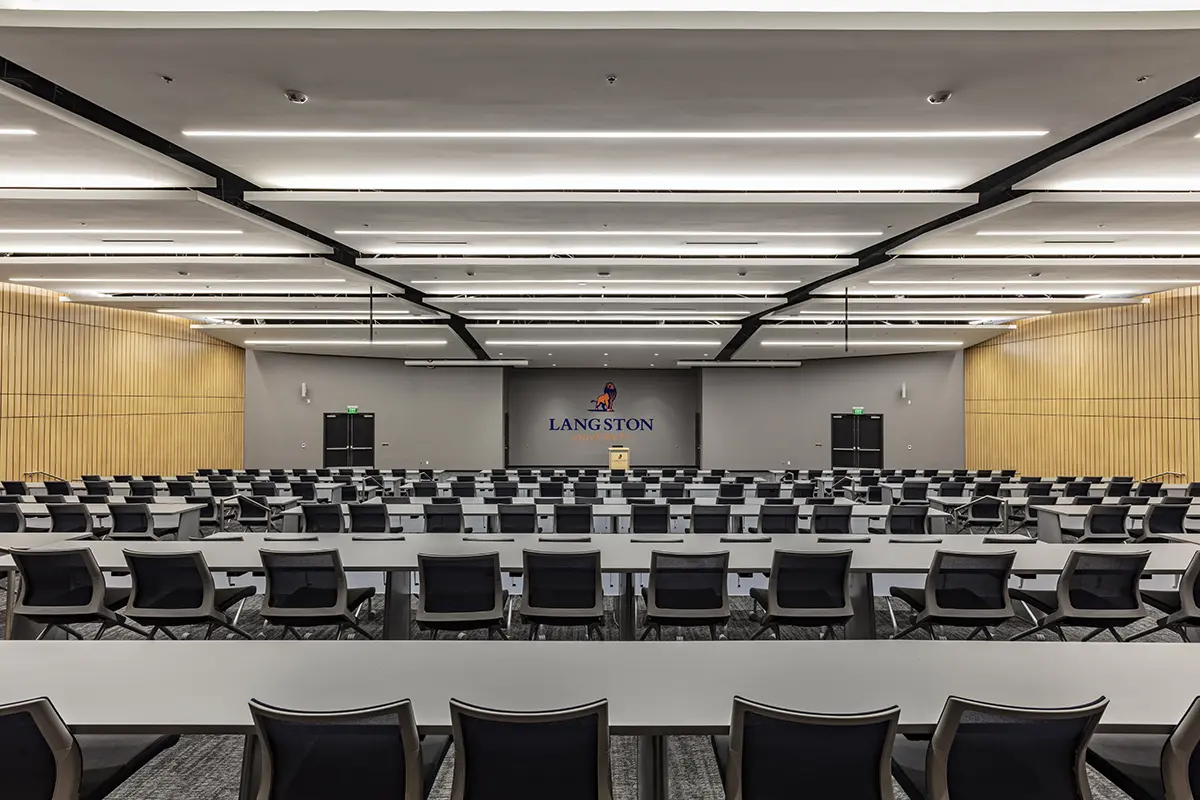 Modern conference room with rows of black chairs and white tables facing a podium and screen at the front. The walls are wooden, and the space is well-lit.
