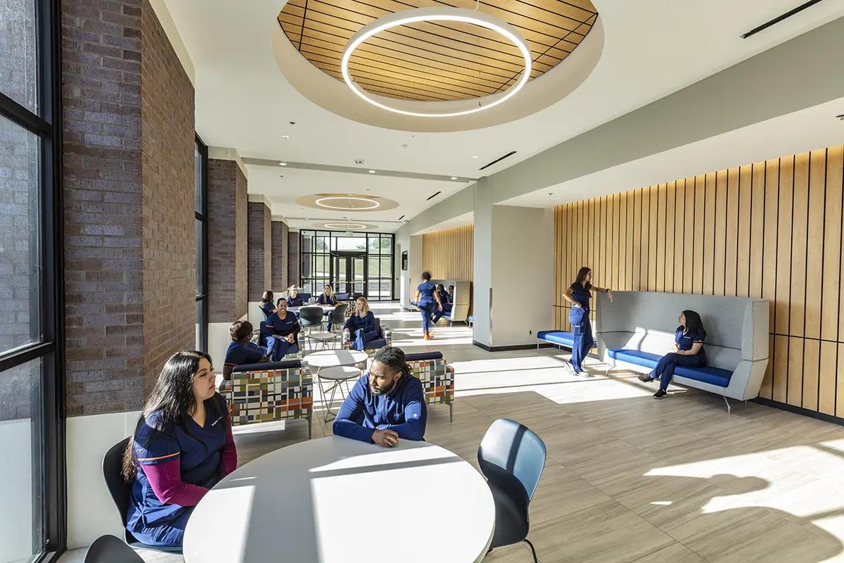 Bright, spacious lounge with large windows and wooden panels. People in blue scrubs sit and chat at tables, conveying a relaxed, collegial atmosphere.