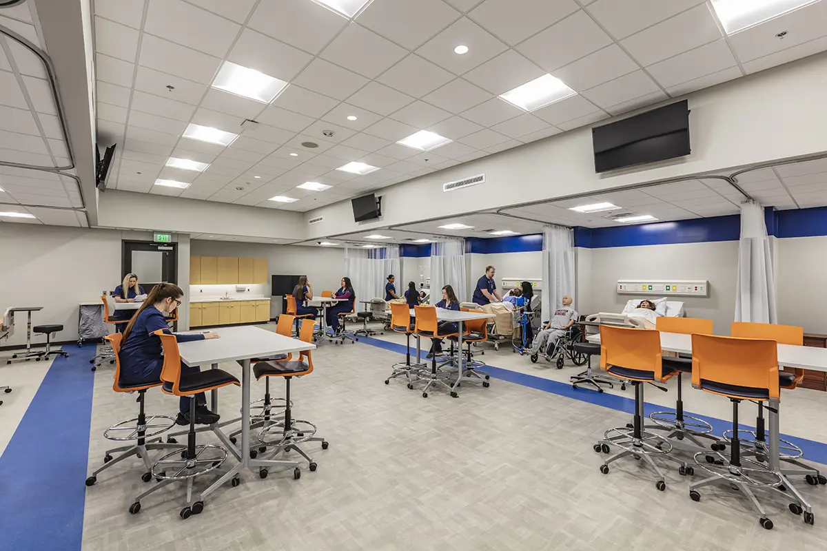 Modern medical training room with partitions, students in blue scrubs at workstations with orange chairs, and instructors by medical mannequins. Brightly lit.
