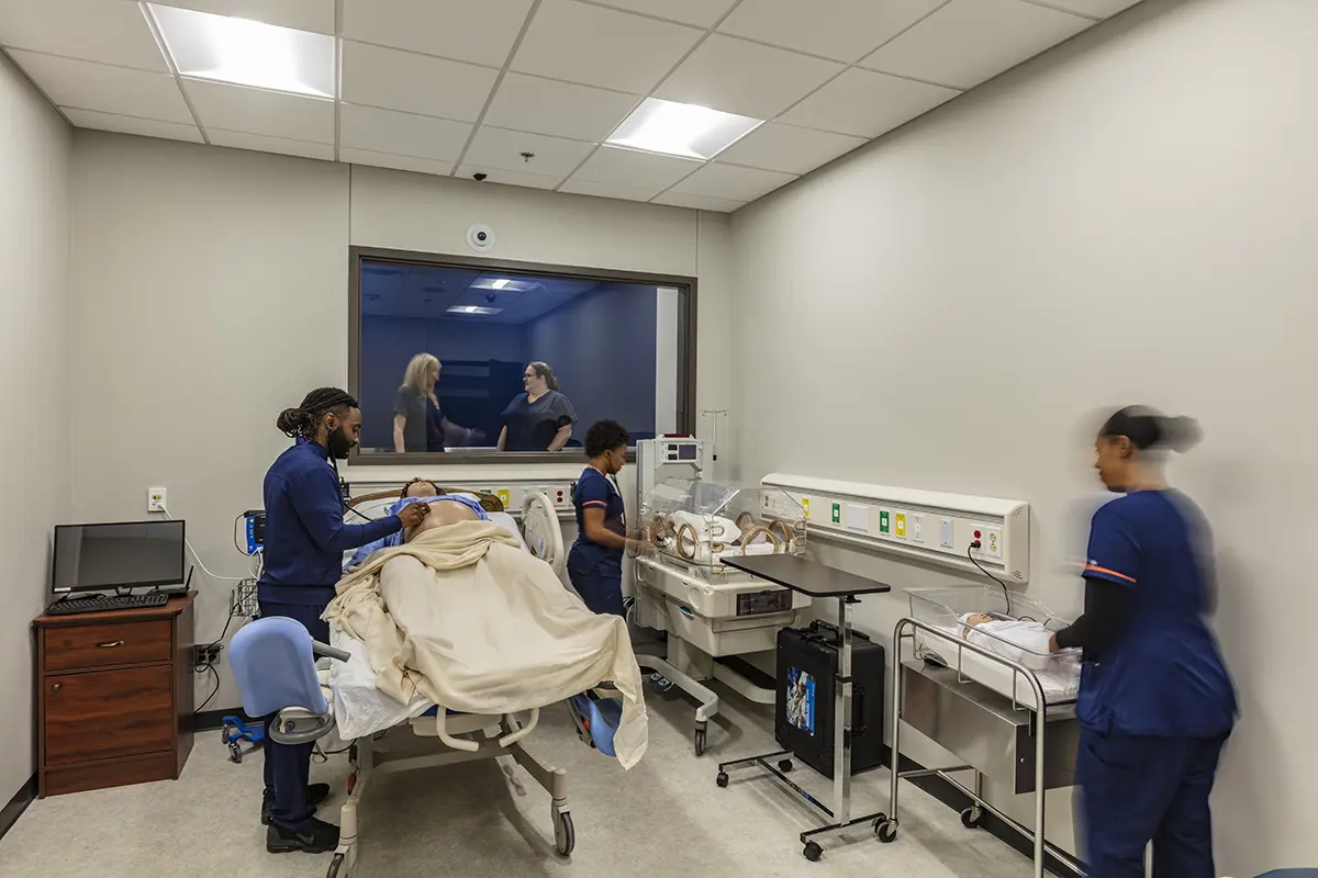 Three medical professionals attend to a patient in a hospital room. Two people observe through a window, creating a focused, collaborative atmosphere.