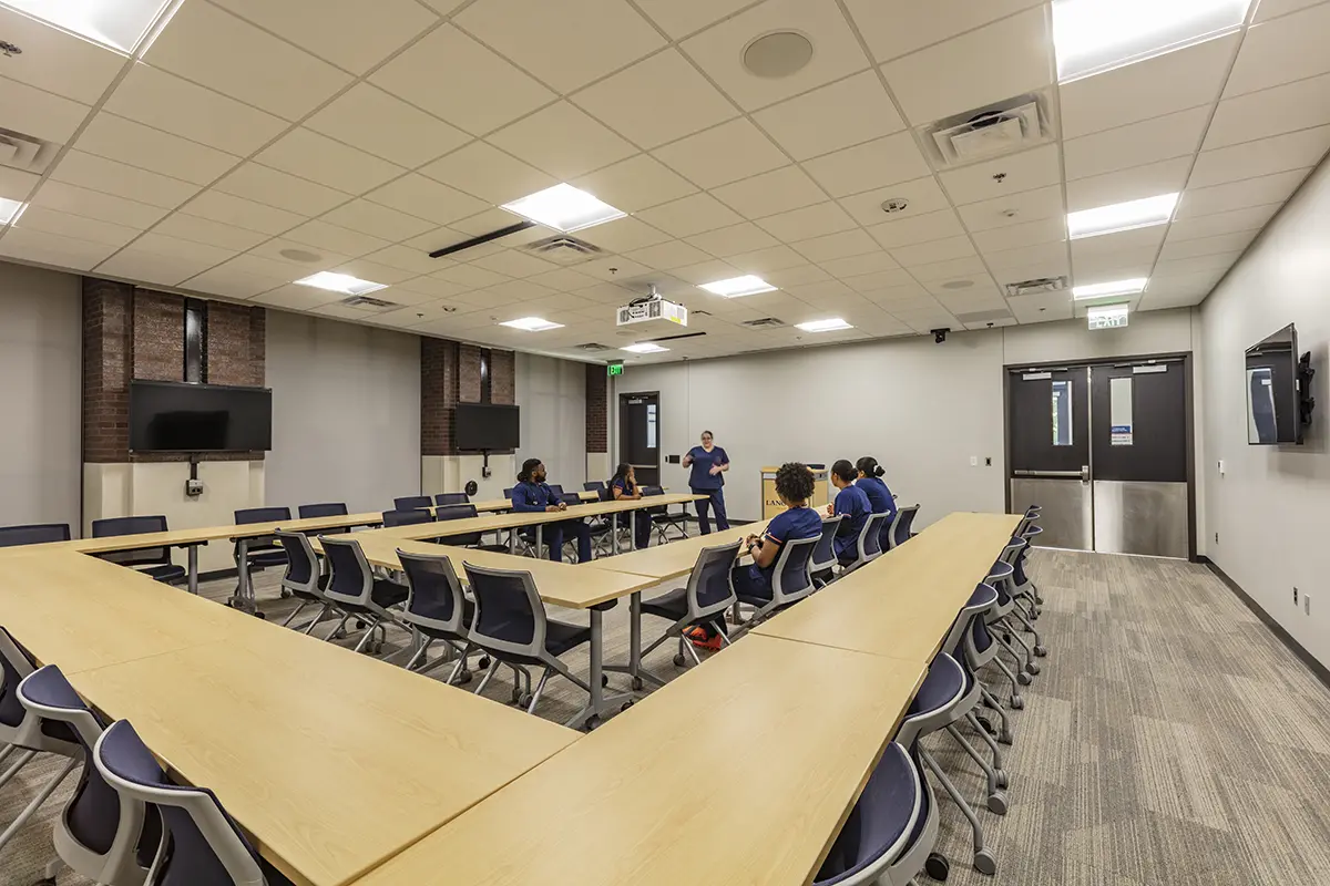 A spacious classroom features a U-shaped table setup with six people in discussion. The room has neutral walls, mounted screens, and a calm atmosphere.