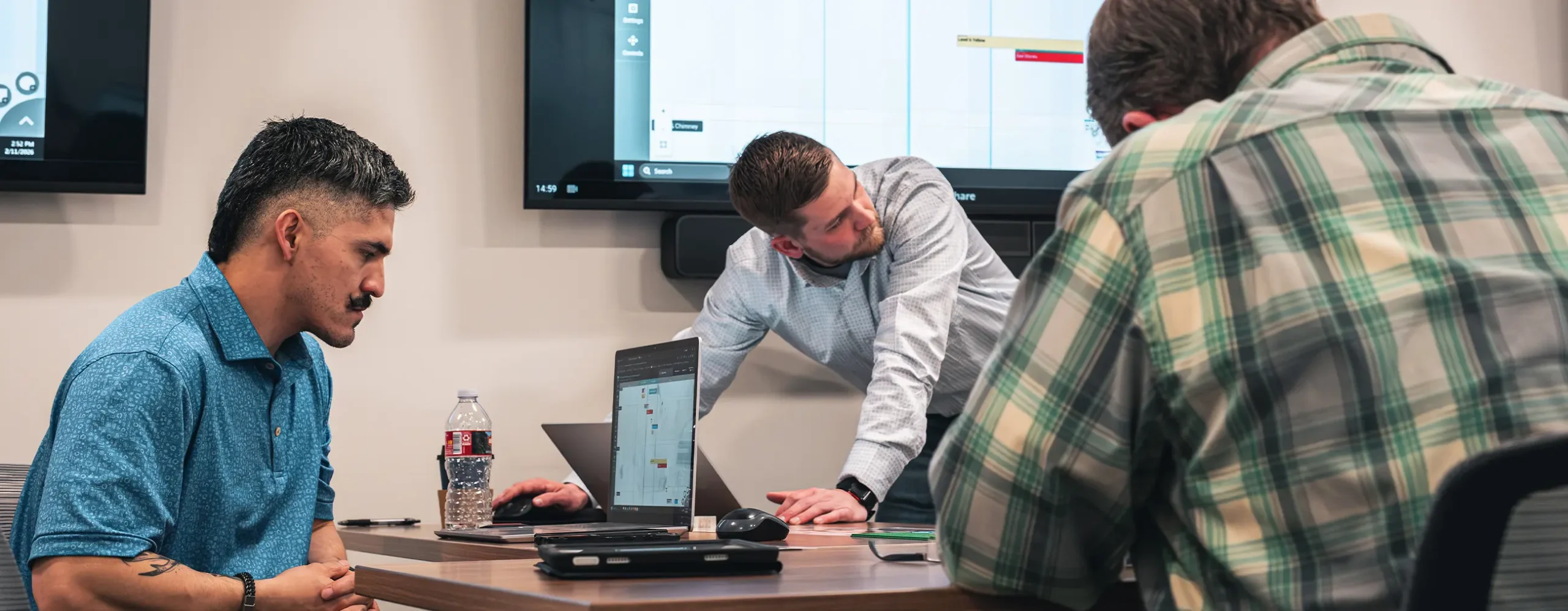 Three men are focused on a meeting in a conference room. Two screens display charts, and a laptop is open on the table, creating a collaborative atmosphere.