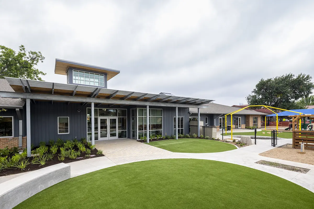 Modern building with a dark exterior and large windows, set in a landscaped area with artificial grass. Nearby, a playground with colorful equipment. Overcast sky.