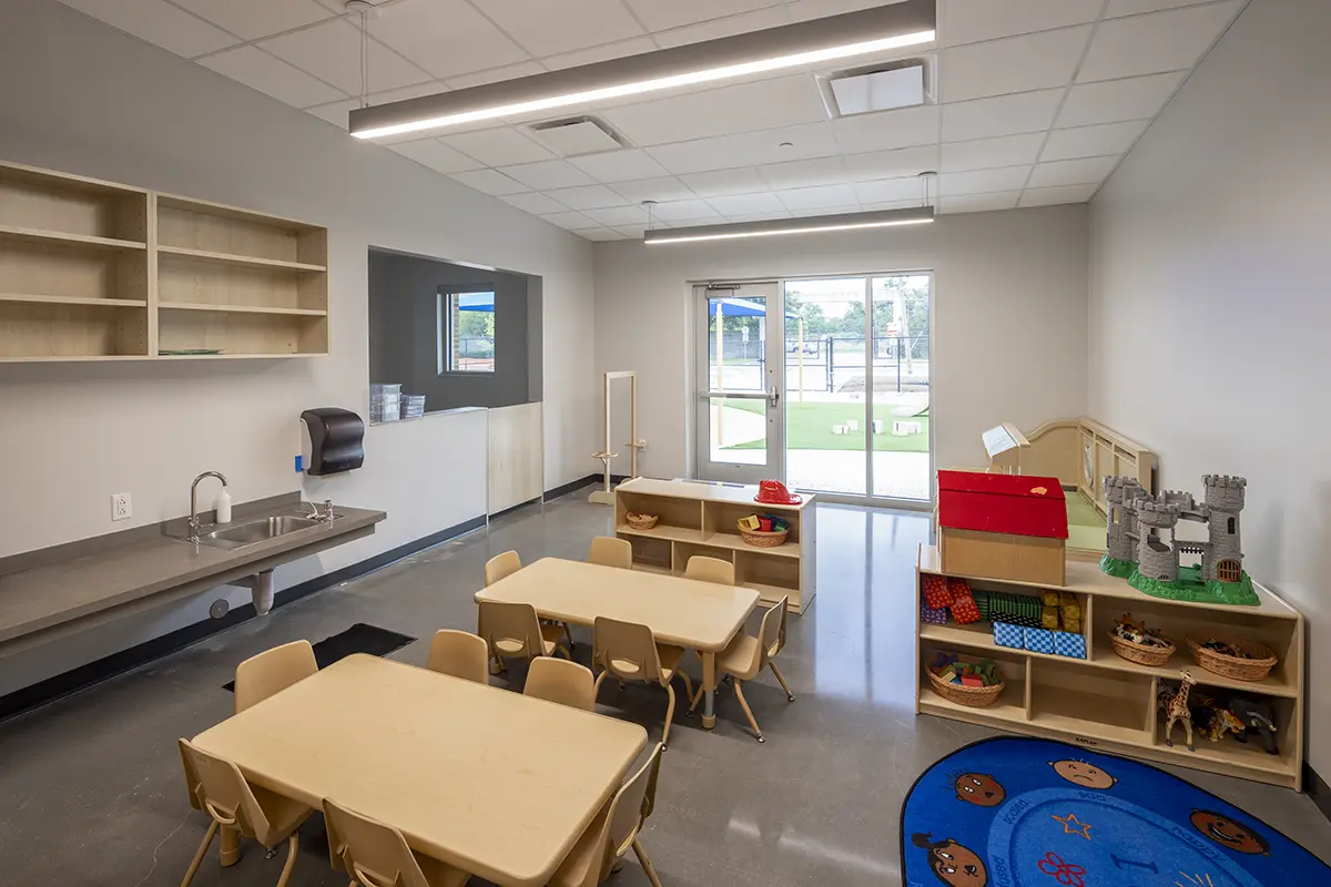 Bright preschool classroom with small wooden tables and chairs, colorful mats, toy shelves, and a play kitchen. Sunlight streams through the large window.
