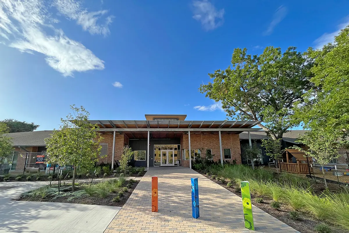 Modern brick building under a clear blue sky, surrounded by greenery. Vibrant red, blue, and green posts line the entrance path, creating a welcoming atmosphere.