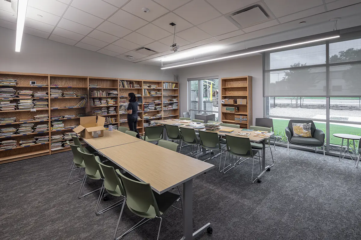 A modern library room with wooden bookshelves filled with books along two walls. A person stands near the shelves. A long table with green chairs is centered, with more books atop. Large windows let in natural light, creating a calm atmosphere.