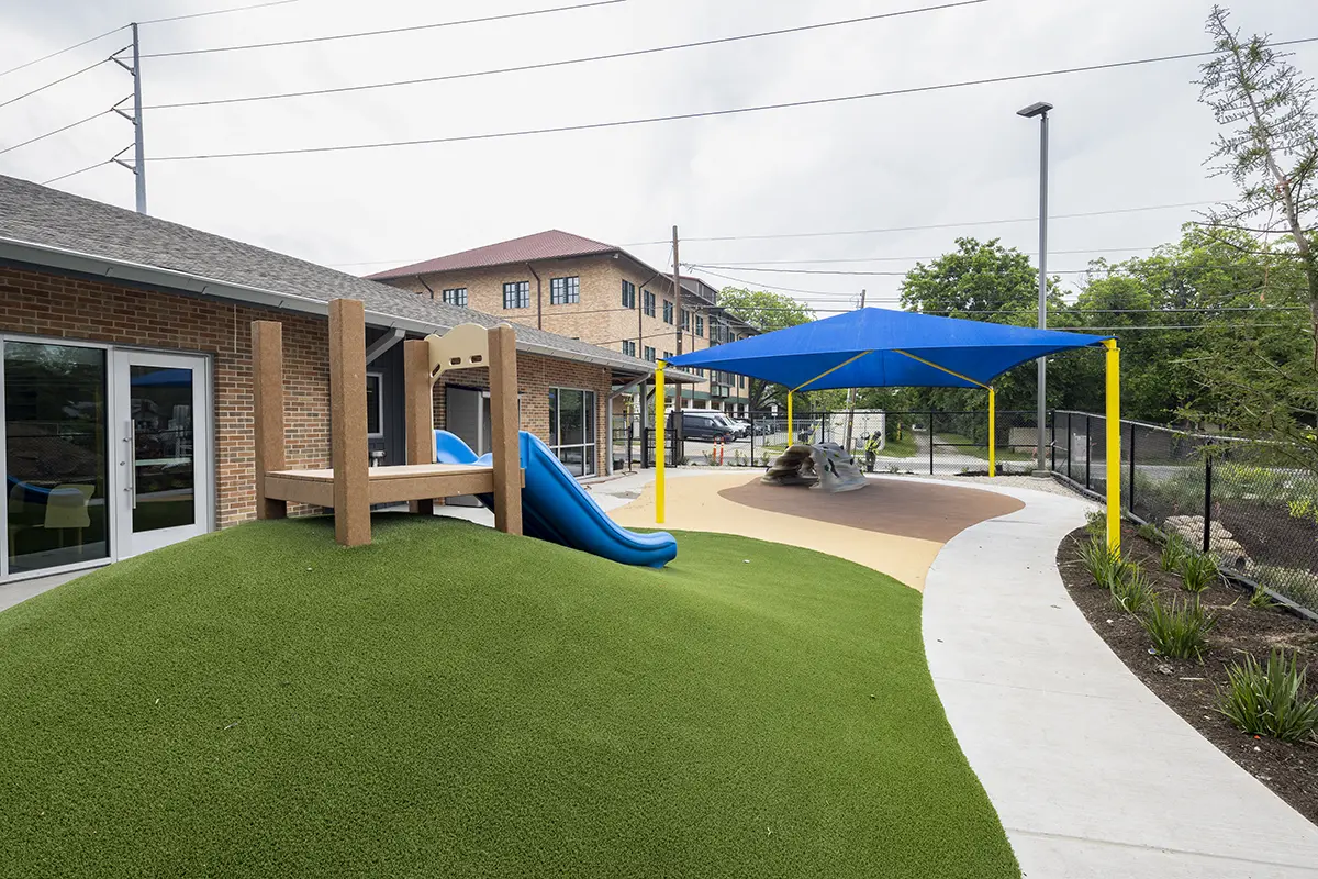 Playground with green artificial hill, blue slide, and large blue canopy. Nearby buildings, concrete paths, and lush greenery create a calm atmosphere.
