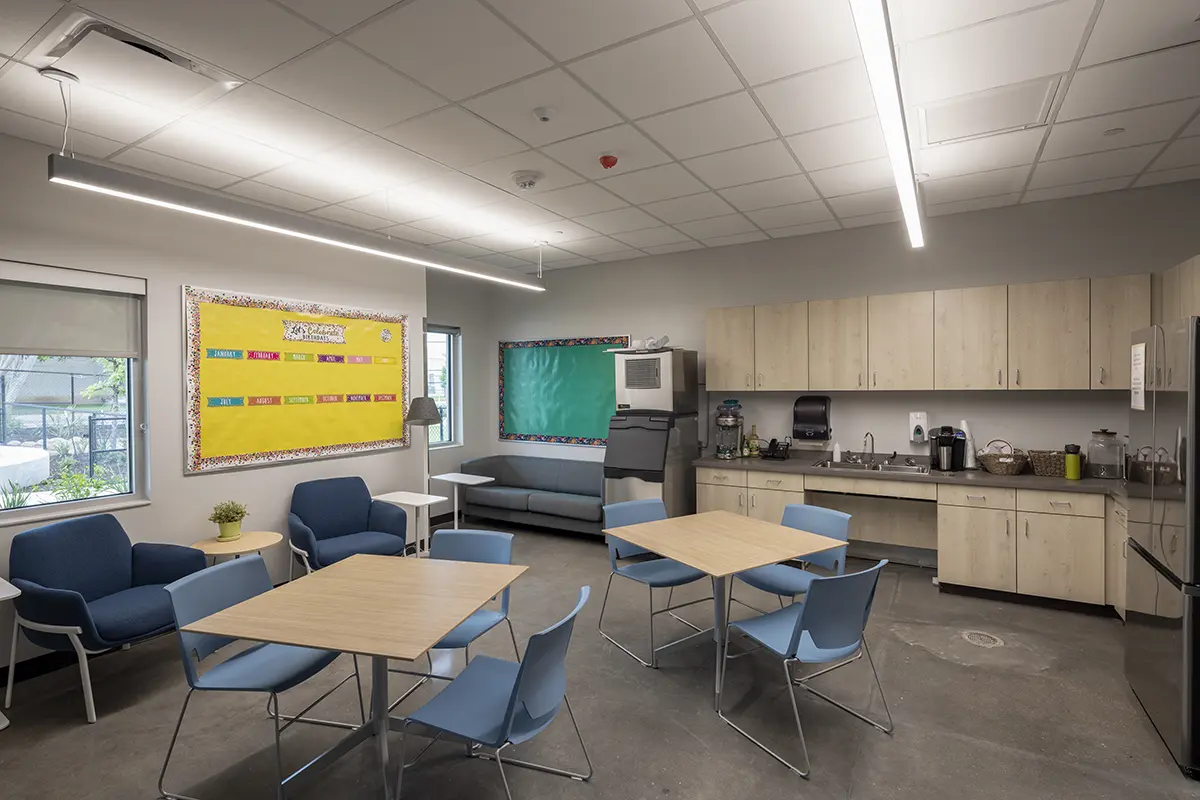 Spacious break room with light wood cabinets, three tables, blue chairs, and cozy seating area. Bulletin boards add a welcoming, organized touch.