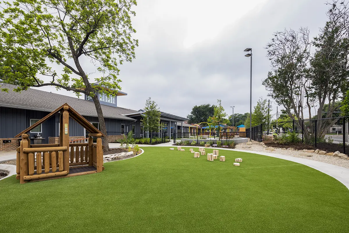 Playground with green artificial turf, wooden playhouse, and scattered wooden seats. Trees and modern building in background under cloudy sky. Calm atmosphere.