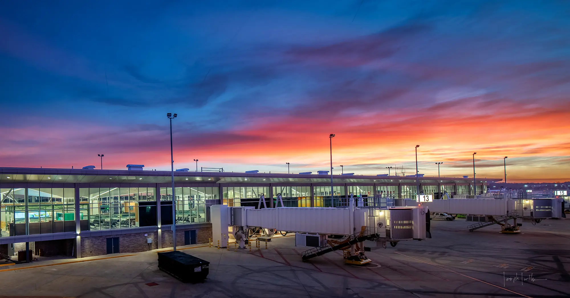 An airport terminal at sunset, featuring a vivid sky in shades of blue, pink, and orange. Jet bridges extend to the tarmac, and the scene is calm and serene.