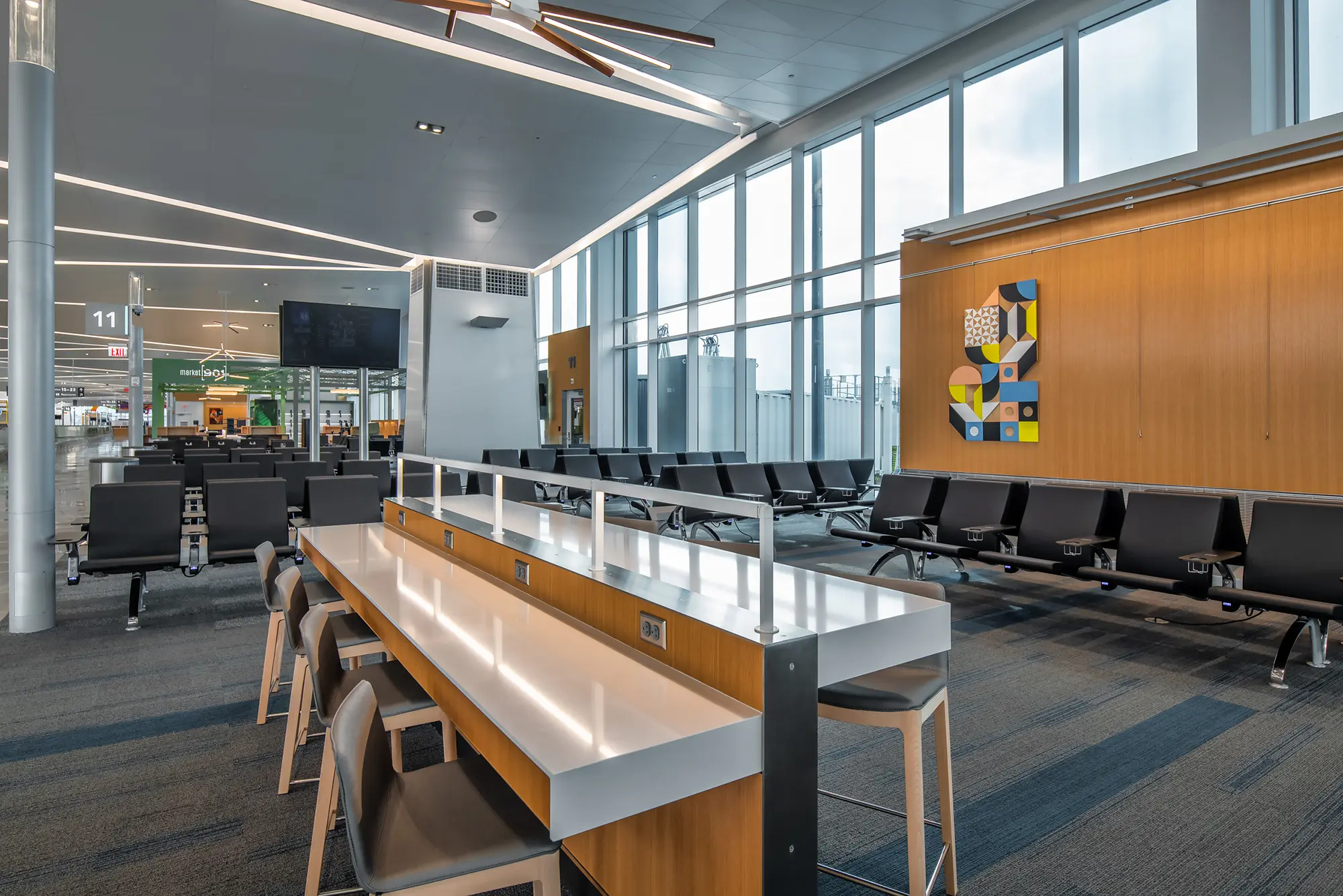 Spacious airport terminal with modern seating and workstations, large windows providing natural light, and a colorful abstract wall art. Quiet, clean ambiance.
