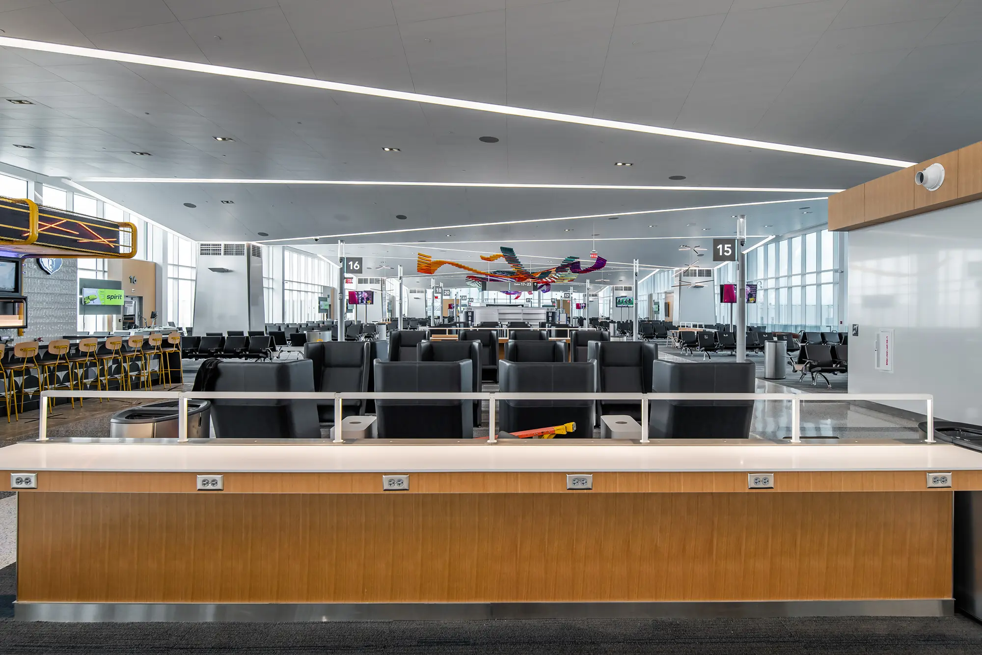 Modern airport gate area with sleek black seating, high ceilings, and bright lighting. A colorful abstract sculpture hangs overhead, creating a vibrant focal point.