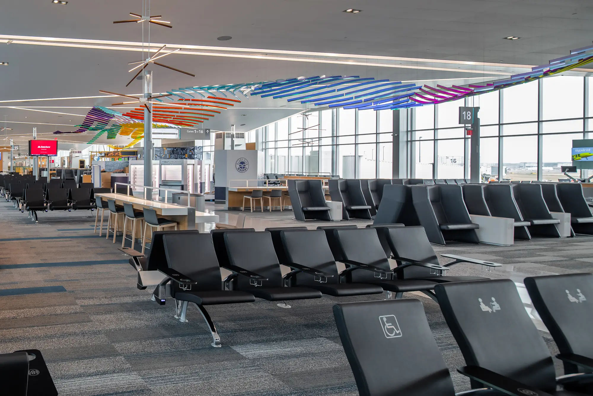 Spacious airport departure lounge with rows of black seating and colorful ceiling art. Large windows fill the area with natural light, creating a calm atmosphere.