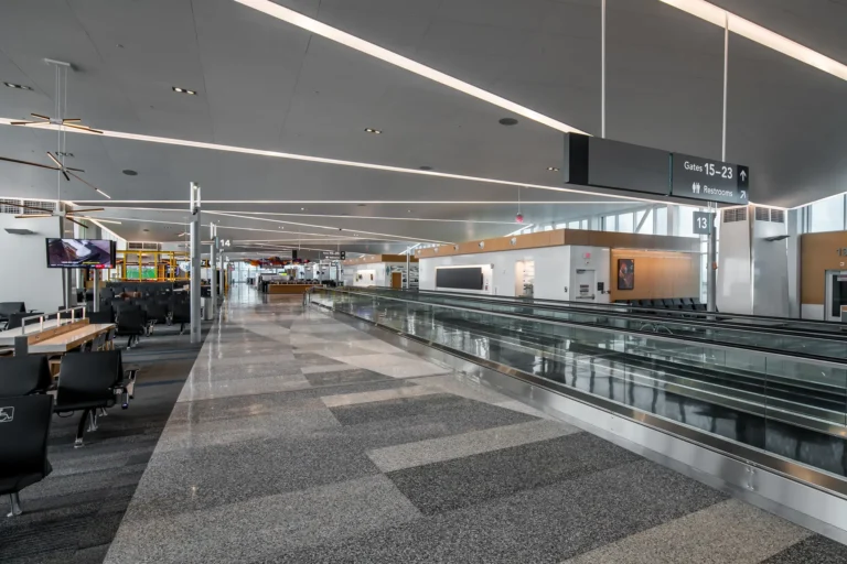 Spacious airport terminal with a glossy floor, empty modern seating area, and an illuminated walkway. Overhead signs guide travelers to gates and restrooms.