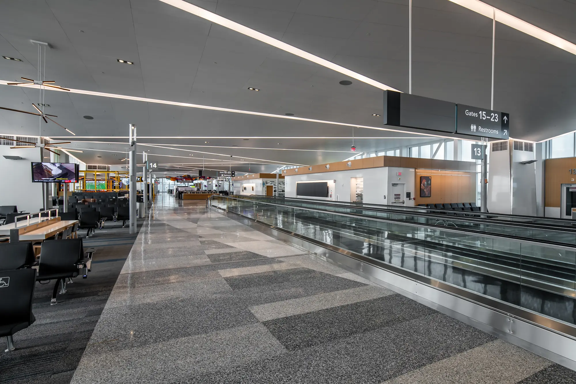 Spacious airport terminal with a glossy floor, empty modern seating area, and an illuminated walkway. Overhead signs guide travelers to gates and restrooms.